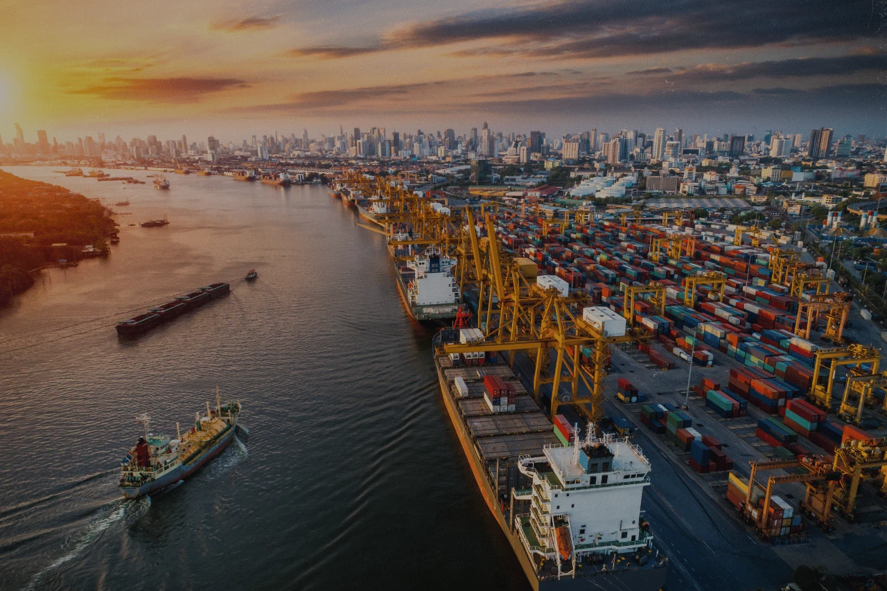 Cargo ships and containers at a busy port with city skyline at sunset.